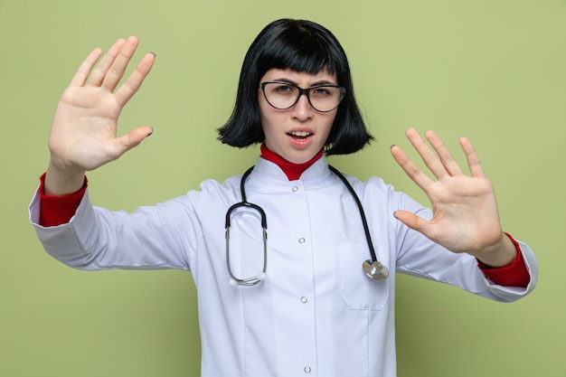 Poważna młoda ładna kaukaska dziewczyna w okularach optycznych w mundurze lekarza ze stetoskopem wskazującym znak stop dwiema rękami