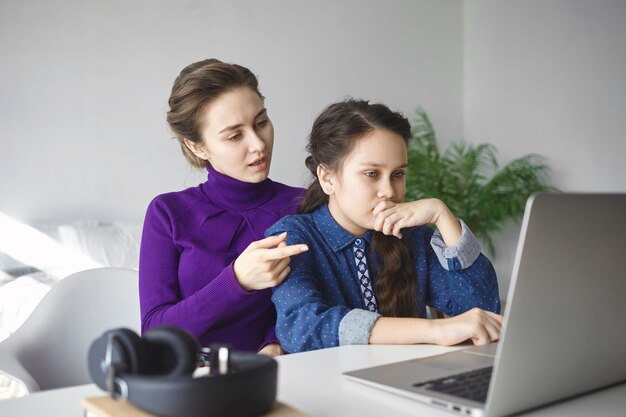 Poważna nastolatka odrabia lekcje, szuka informacji w internecie za pomocą laptopa, podczas gdy matka jej pomaga,