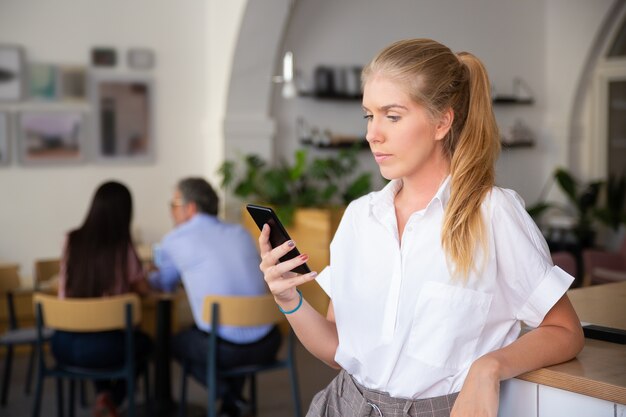 Poważna piękna młoda kobieta ubrana w białą koszulę, za pomocą smartfona, wpisując wiadomość, stojąc w przestrzeni coworkingowej