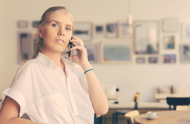 Bezpłatne zdjęcie poważna zamyślona piękna młoda kobieta ubrana w białą koszulę, rozmawiająca przez telefon komórkowy, stojąca w przestrzeni coworkingowej