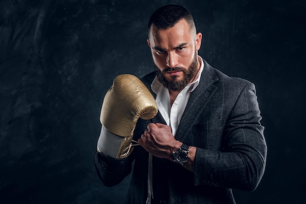 Poważny brutalny mężczyzna w garniturze i złotej rękawicy bokserskiej pozuje dla fotografa w ciemnym studiu fotograficznym.