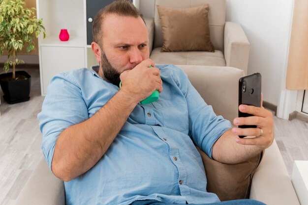 Poważny dorosły słowiański mężczyzna siedzi na fotelu, pijąc z kubka i patrząc na telefon w salonie