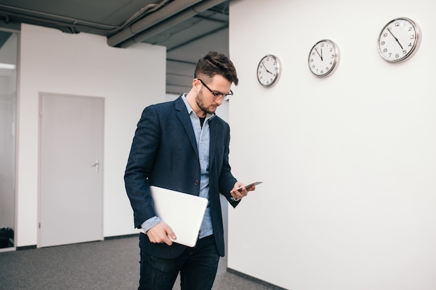 Poważny facet w okularach chodzi w biurze. Nosi niebieską koszulę, ciemną kurtkę, dżinsy i brodę. On pisze na telefonie i trzyma w ręku laptopa.