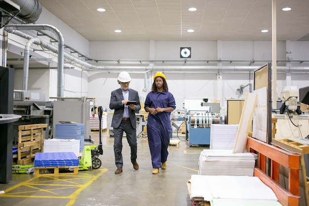 Poważny inspektor płci męskiej i pracownica fabryki w kaskach chodzą po hali produkcyjnej i rozmawiają, mężczyzna za pomocą tabletu