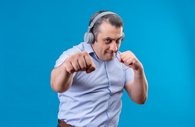Poważny mężczyzna w średnim wieku ubrany w niebieską koszulę w pionowe paski w słuchawkach ćwiczy ruchy bokserskie na niebieskim tle