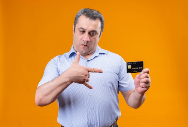 Poważny mężczyzna w średnim wieku w niebieskiej koszuli w paski, patrząc na kartę kredytową na pomarańczowym tle