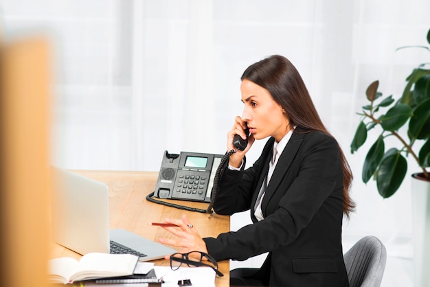 Bezpłatne zdjęcie poważny młody bizneswomanu obsiadanie przy biurkiem opowiada na telefonie