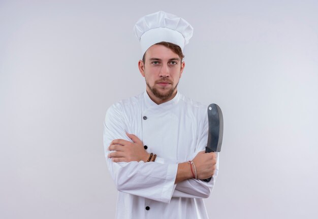 Poważny młody brodaty szef kuchni ubrany w biały mundur kuchenki i kapelusz trzymając tasak do mięsa, patrząc na białej ścianie