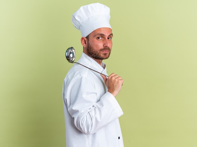 Poważny młody kaukaski kucharz w mundurze szefa kuchni i czapce stojącej w widoku profilu trzymając chochlę na ramieniu, patrząc na kamerę odizolowaną na oliwkowozielonej ścianie z kopią przestrzeni