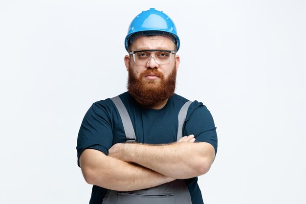 Poważny młody mężczyzna robotnik budowlany noszący mundur w kasku ochronnym i okulary ochronne, patrzący na kamerę ze skrzyżowanymi rękami na białym tle