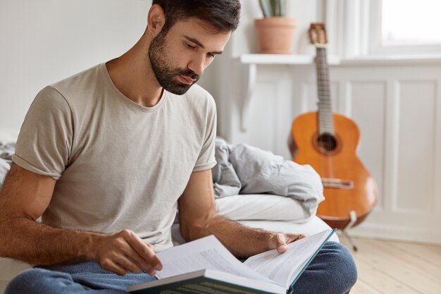 Poważny młody właściciel przedsiębiorczości, studiuje literaturę biznesową, ubrany w luźną koszulkę, odpoczywa na łóżku w swoim pokoju, gitara akustyczna stoi w ścianie. Ludzie, dom, studia, koncepcja czytania