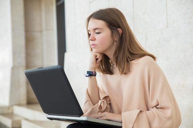 Poważny żeński nastolatek wyszukuje na laptopie na ławce przy budynkiem