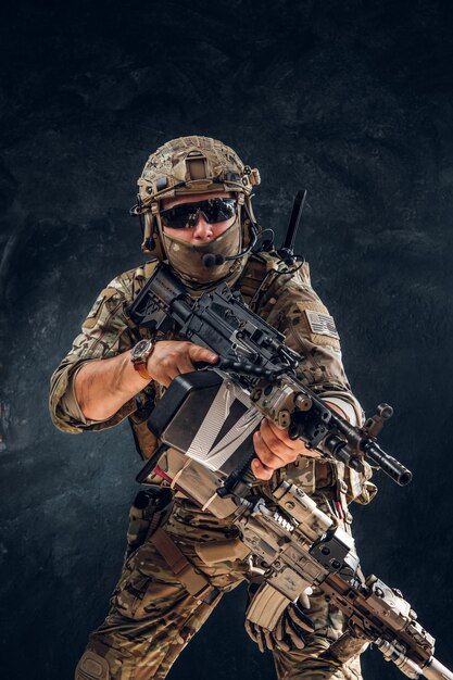 Poważny żołnierz w pełnym ekwipunku i mundurze wojskowym stoi z karabinem maszynowym na ciemnym tle.