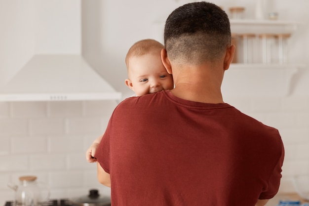 Powrót widok portret m?odych doros?ych Caucasion ojca gospodarstwa syna lub córk?, cute baby patrz?c na kamery na ramieniu mans, stwarzaj?ce kryty z kuchni ustawi? na tle.