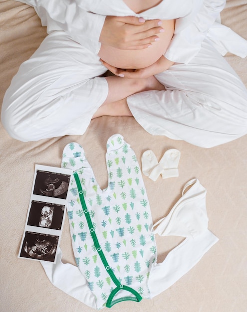 Powyżej Widok Anonimowej Kobiety W Białej Przytulnej Piżamie Siedzącej Na łóżku Z Obrazem Usg Przedstawiającym łono śliczne Body I Skarpetki, Pieszczące Ciężarny Brzuch I Oczekujące Na Dziecko I Część Rodziny