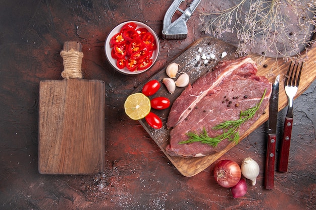 Powyżej widok czerwonego mięsa na drewnianej desce do krojenia i czosnku, zielonej cytryny, cebuli, widelca i noża na ciemnym tle