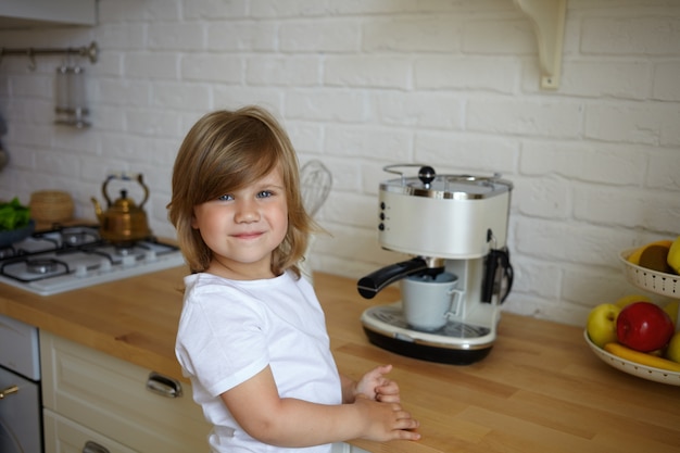 Bezpłatne zdjęcie poziome strzał słodkie beztroskie dziecko płci żeńskiej w wieku przedszkolnym na sobie białą koszulkę o szczęśliwym wyglądzie, stojąc przy kuchennym stole, robiąc kawę dla jej ojca. beztroskie dzieciństwo i koncepcja gotowania