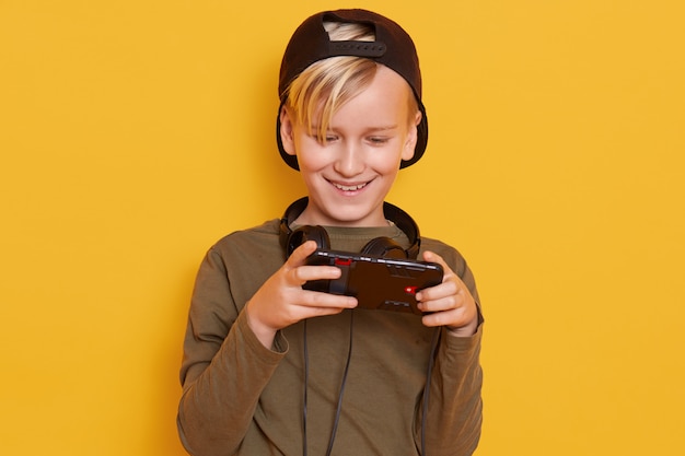 Poziome ujęcie małego chłopca w czarnej czapce i zielonej bluzie z kapturem, pozującego z telefonem komórkowym w rękach, modnego dzieciaka grającego w gry online.