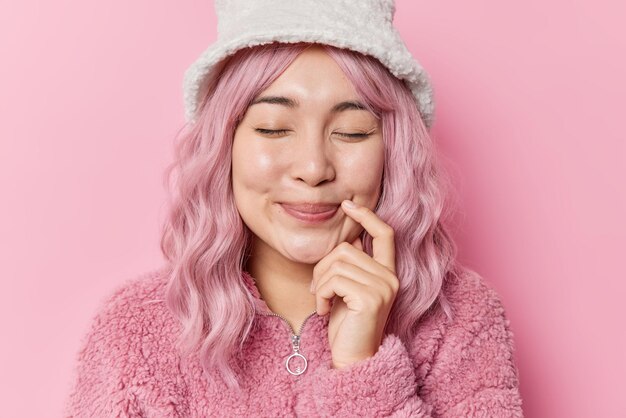 Poziome ujęcie rozmarzonej zachwyconej Azjatki zamyka oczy i trzyma palec blisko kącika ust ma różowo farbowane włosy ubrane w futro i zimowy kapelusz na białym tle nad różowym tłem Szczęśliwe uczucia