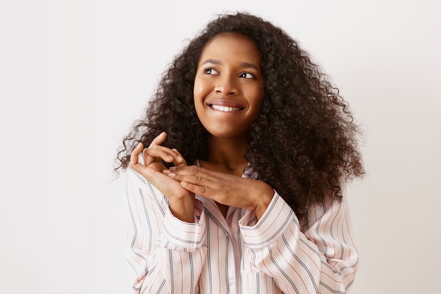 Poziome Ujęcie Uroczej Młodej Afro Amerykanki W Stylowej Koszuli Nocnej, Patrzącej W Górę Z Podekscytowanym Zamyślonym Wyrazem Twarzy, Gryzącej Wargę I Zacierającej Ręce, Mającej Genialny Pomysł Lub Plan, śniącej