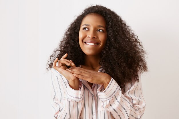 Poziome ujęcie uroczej młodej Afro Amerykanki w stylowej koszuli nocnej, patrzącej w górę z podekscytowanym zamyślonym wyrazem twarzy, gryzącej wargę i zacierającej ręce, mającej genialny pomysł lub plan, śniącej