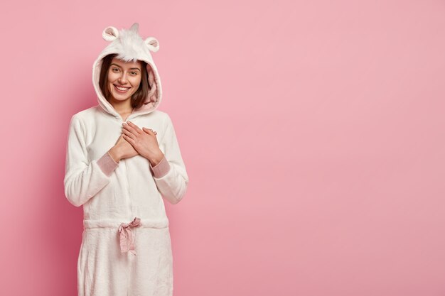 Poziome ujęcie uśmiechniętej młodej kobiety ma pozytywny uśmiech, trzyma ręce na piersi, wyraża szczere emocje, nosi biały kostium kigurumi z uszami, stoi nad różową ścianą z pustą przestrzenią