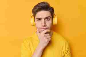 Bezpłatne zdjęcie poziome ujęcie zamyślonego mężczyzny trzymającego podbródek i patrzącego poważnie w kamerę, słuchającego ulubionej piosenki przez słuchawki stereo, ubranego w zwykłe ubrania pozuje na żywym żółtym tle niech pomyślę