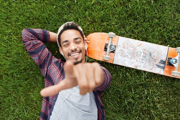 Poziomy portret radosnego, brodatego mężczyzny skater leży na zielonej trawie w pobliżu deskorolki, słucha muzyki przez słuchawki