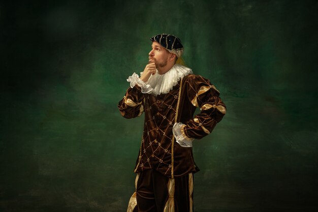 Pozowanie w zamyśleniu. Portret średniowiecznego młodzieńca w odzież vintage, stojąc na ciemnym tle. Męski model jako książę, książę, osoba królewska. Pojęcie porównania epok, nowoczesności, mody.