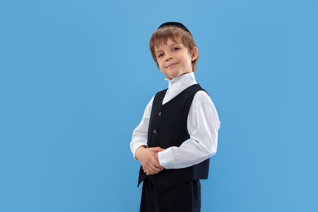 Pozuje pewny siebie, słodki. portret młodego ortodoksyjnego żydowskiego chłopca na białym tle na niebieskiej ścianie. purim, biznes, festiwal, wakacje, obchody pesach lub pascha, judaizm, koncepcja religii.