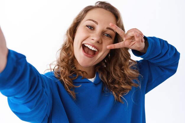 Pozytywna atrakcyjna dziewczyna biorąca selfie, pokazująca gest pokoju znak v w pobliżu oka i trzymająca aparat z wyciągniętą ręką, robiąca zdjęcie na smartfonie, biała ściana