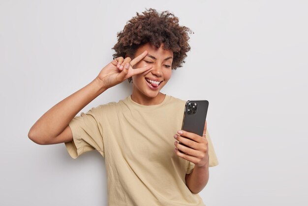 Pozytywna atrakcyjna kobieta z kręconymi włosami sprawia, że gest pokoju nad okiem wystaje język pozy do selfie na ekranie smartfona, ubrana w casualową beżową koszulkę na białym tle nad białym