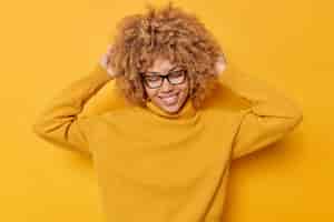 Bezpłatne zdjęcie pozytywna beztroska kobieta dobrze się bawi trzyma ręce na głowie uśmiech delikatnie zamyka oczy z zadowolenia nosi okulary a sweter z długim rękawem przypomina coś ładnego odizolowanego na żółtym tle