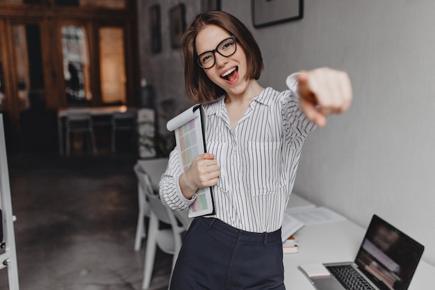 Bezpłatne zdjęcie pozytywna biznesowa kobieta uśmiecha się i wskazuje palcem na aparat. kobieta w spodniach i bluzce pozuje z dokumentami w tle pakietu office.