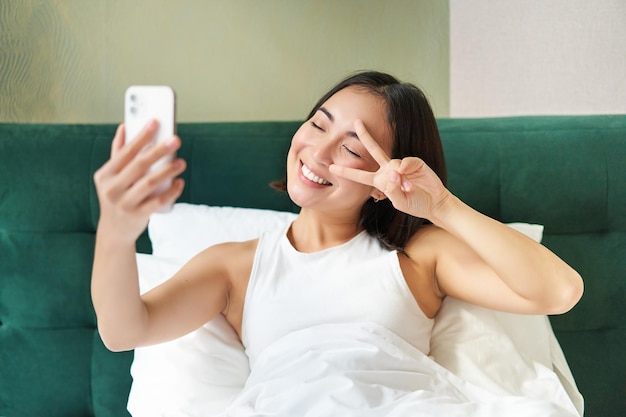 Pozytywna Blogerka Robi Selfie Na Zdjęciu W Sypialni Na Smartfonie, Uśmiechając Się I Pokazując Spokój