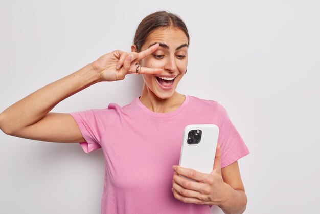 Pozytywna brunetka Europejka robi gest pokoju nad okiem, trzymając telefon komórkowy i robi selfie sukienki w casualowej różowej koszulce pozuje na białym tle Ludzie zabawy i koncepcja technologii