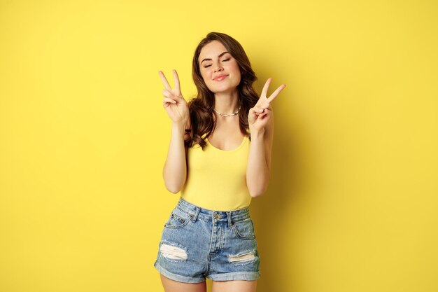 Pozytywna dziewczyna, modelka pokazująca spokój, gest w kształcie litery v i uśmiechnięta, stojąca w podkoszulku i dżinsowych szortach, żółte tło