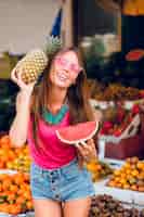 Bezpłatne zdjęcie pozytywna dziewczyna z wielkim uśmiechem trzyma ananas i kawałek arbuza na rynku