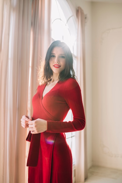 Bezpłatne zdjęcie pozytywna elegancka młoda kobieta w czerwieni sukni blisko okno w lekkim pokoju