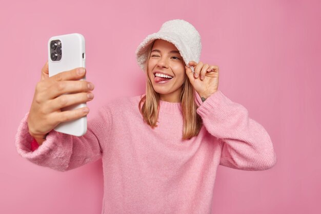 Pozytywna młoda blondynka mruga okiem i wysuwa język, robi selfie lub wideorozmowę przez smartfon, ma na sobie nowy luźny sweter panama na różowym tle, głupcy wokół mają optymistyczny nastrój