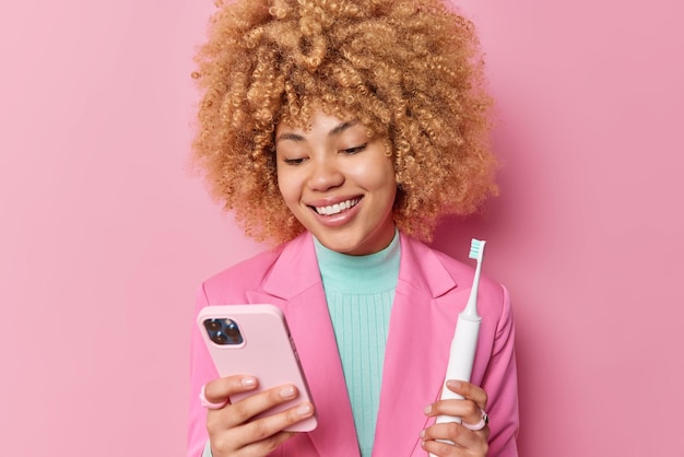 Pozytywna młoda kędzierzawa kobieta ubrana w elegancką kurtkę trzyma telefon komórkowy i elektryczną szczoteczkę do zębów poddawanych procedurom higienicznym, sprawdza powiadomienie otrzymane na komórce odizolowanej na różowym tle