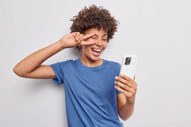 Pozytywna młoda kobieta robi gest pokoju nad okiem robi selfie za pomocą przedniej kamery smartfona, uśmiecha się szeroko ubrana w casualową niebieską koszulkę na białym tle na białym tle ma beztroski wyraz