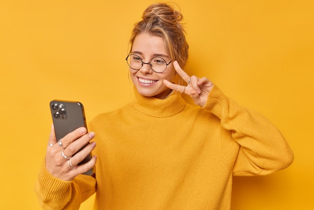 Pozytywna młoda kobieta sprawia, że gest pokoju bierze selfie robi zdjęcie siebie ubranej w luźne okrągłe okulary na białym tle na żółtym tle. Hej obserwatorzy, jak mija dzisiaj dzień