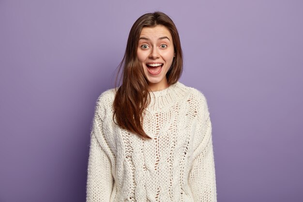 Pozytywna młoda kobieta z radosnym wyrazem twarzy, otwiera usta ze zdziwienia, reaguje na pozytywne, niespodziewane wieści, nosi biały sweter, stoi pod fioletową ścianą, zaintrygowana powiedzeniem