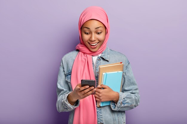 Pozytywna muzułmanka patrzy na urządzenie smatphone, nosi różowy hidżab, dżinsową kurtkę