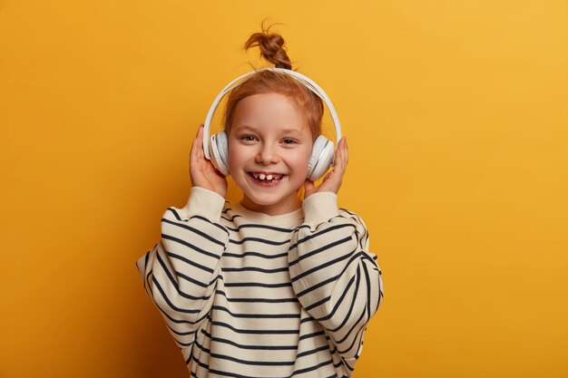 Pozytywna ruda dziewczyna lubi ulubioną melodię, słucha muzyki w słuchawkach, ma optymistyczny nastrój, zawiązane włosy, nosi luźny sweter w paski, pozuje na żółtej ścianie, uśmiecha się zębami