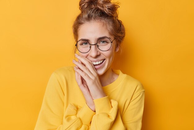 Pozytywna szczęśliwa kobieta śmieje się z radością trzyma ręce przy ustach wyraża autentyczne, szczere emocje nosi okulary i swobodny sweter na białym tle nad żywym żółtym tłem Pojęcie szczęścia
