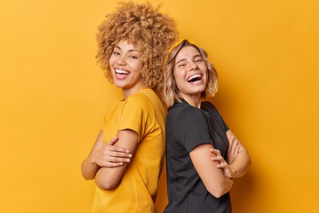 Pozytywne dwie młode ładne kobiety stoją tyłem do siebie, śmieją się z założonymi rękoma, szczęśliwie ubrane w zwykłe koszulki, śmieją się z czegoś zabawnego izolowanego na żółtym tle Koncepcja przyjaźni