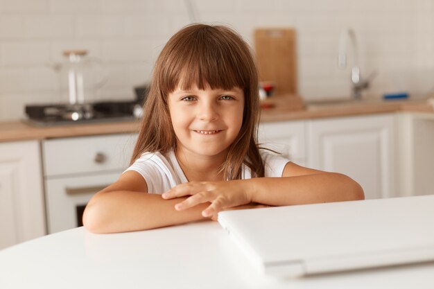 Pozytywne małe ciemnowłose dziecko płci żeńskiej siedzi przy stole w pobliżu złożonego laptopa, patrząc na kamerę z przyjemnym wyrazem twarzy, pozowanie w domu w kuchni.