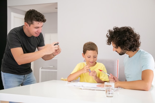 Pozytywne słodkie dziecko robi domowe zadanie szkolne z pomocą dwóch ojców, pisząc w papierach. Człowiek robi zdjęcie swojej rodziny. Koncepcja rodziny i rodziców gejów